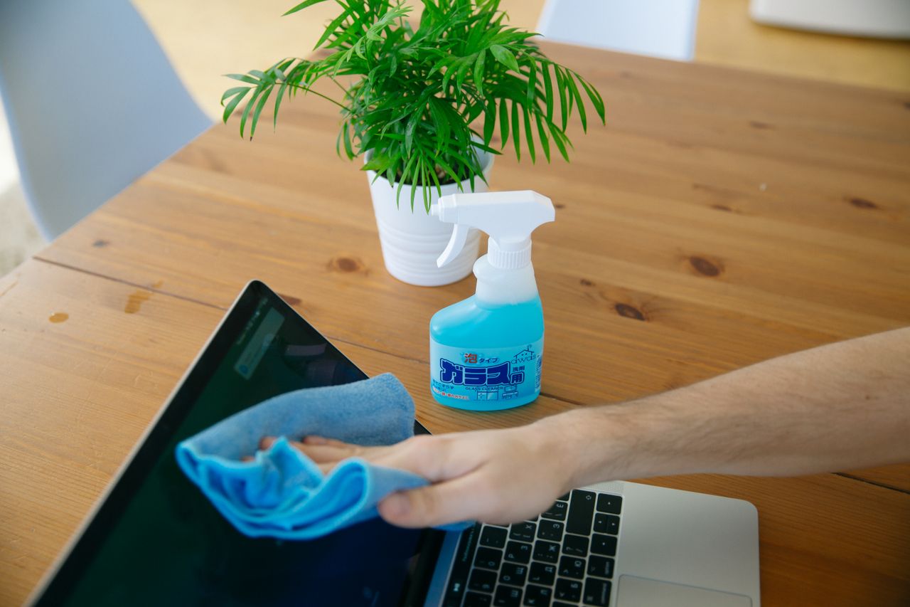 A tight shot of a hand wiping a laptop display with a blue cloth beside a spray bottle on a wooden desk.